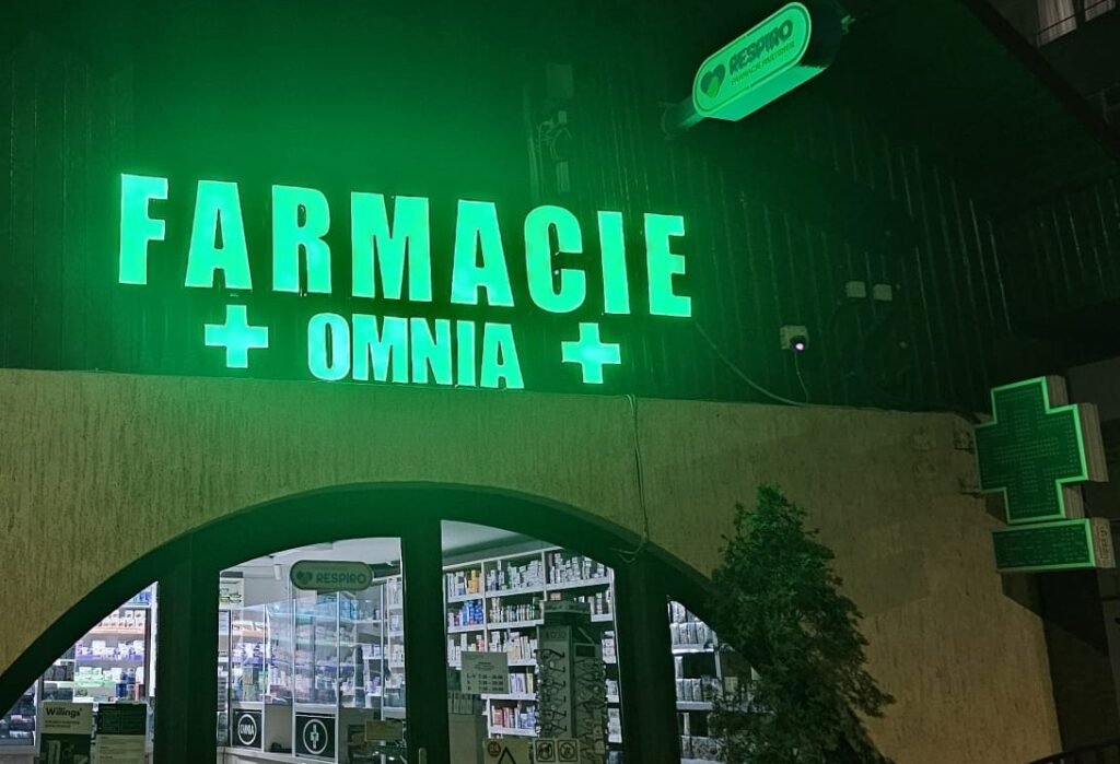 Exteriorul farmaciei Omnia FarmImpex iluminat pe timp de noapte, cu firmă luminoasă verde și intrare modernă, reflectând profesionalism, încredere și tradiție.